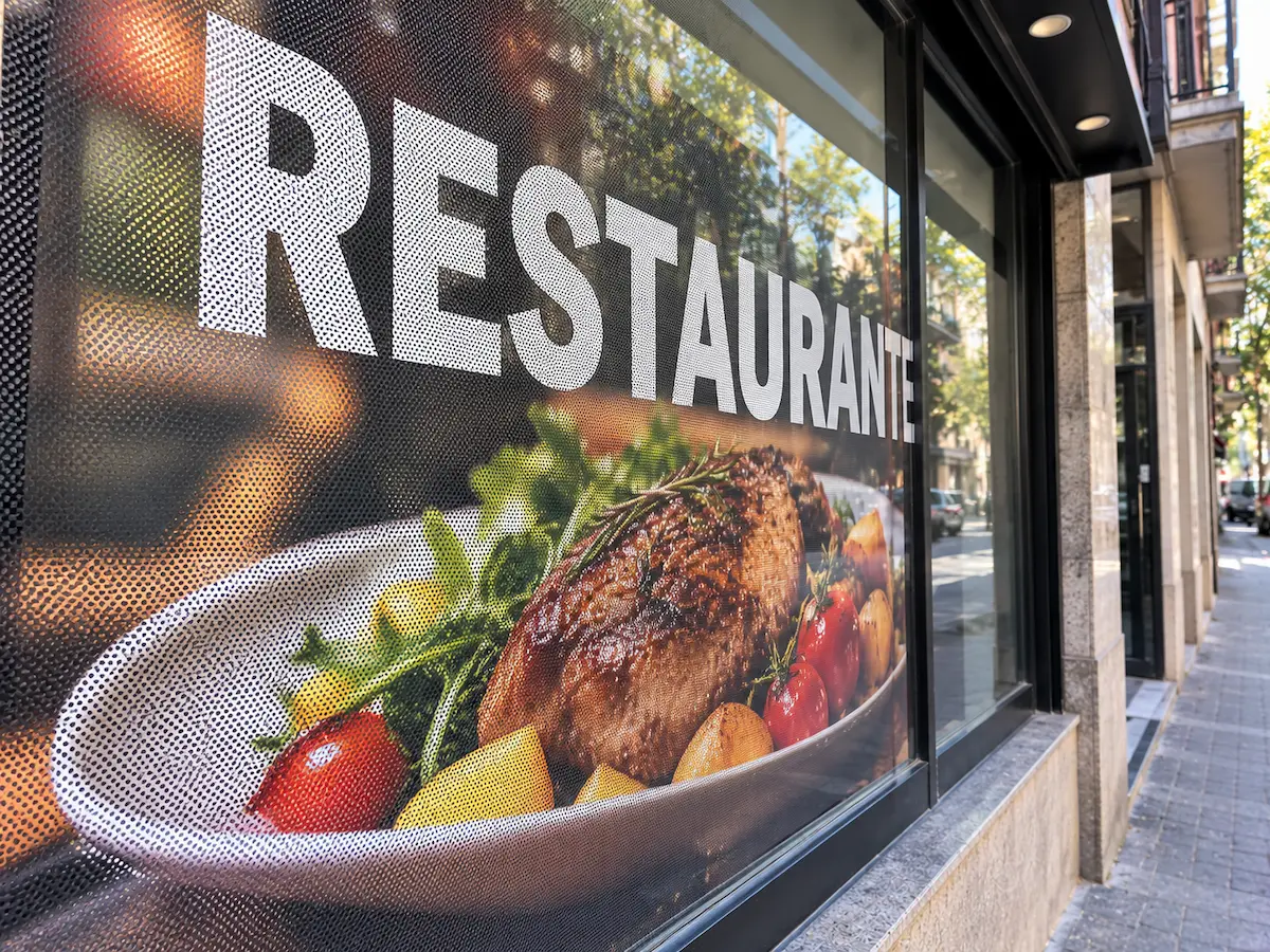 Escaparate visto desde la calle con vinilo microperforado impreso con un plato de carne y verduras y la palabra RESTAURANTE, con la trama de puntos visible